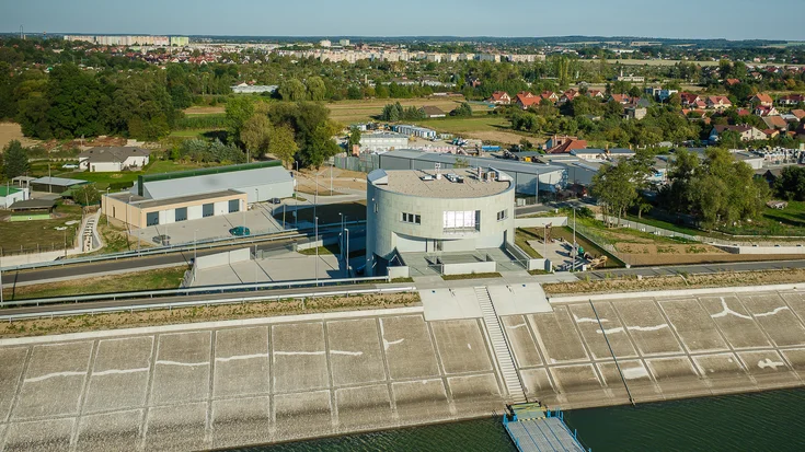 Zylinderförmiges Servicegebäude in der Mitte des Bildes; davor der Stausee in Nysa; dahinter Wald und kleine Häusersiedlung