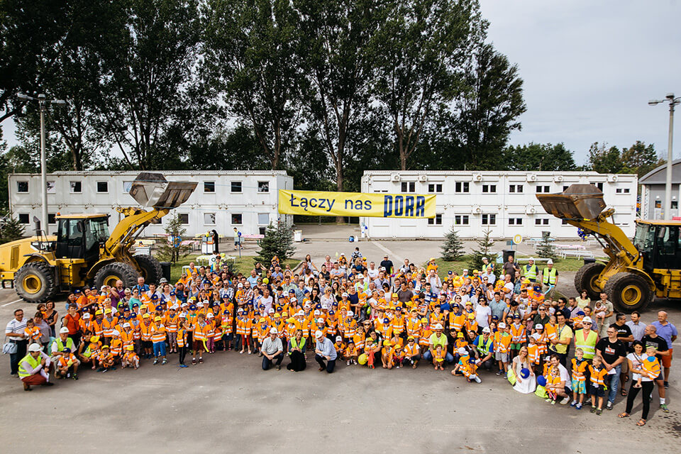 Gruppenfoto mit mehr als 100 Kindern in orangen Warnwesten und ihren Angehörigen, dahinter rechts und links zwei Bagger, die ein Transparent „We are connected by PORR“